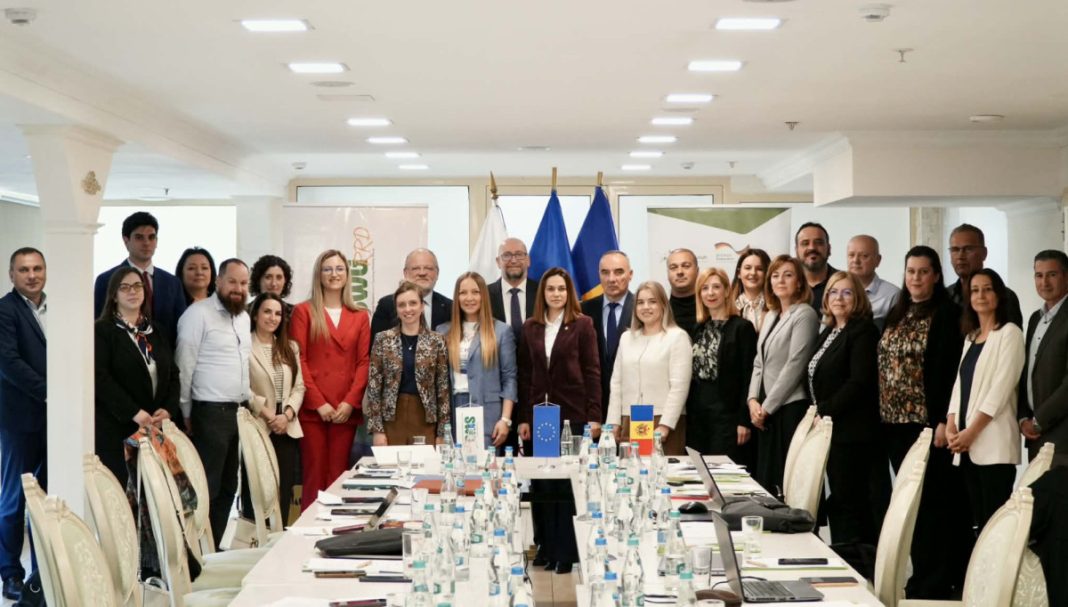 Group of professionals in business attire posing for a formal group photo in a conference room, with flags behind them and a long table set with water bottles, notebooks, and laptops.