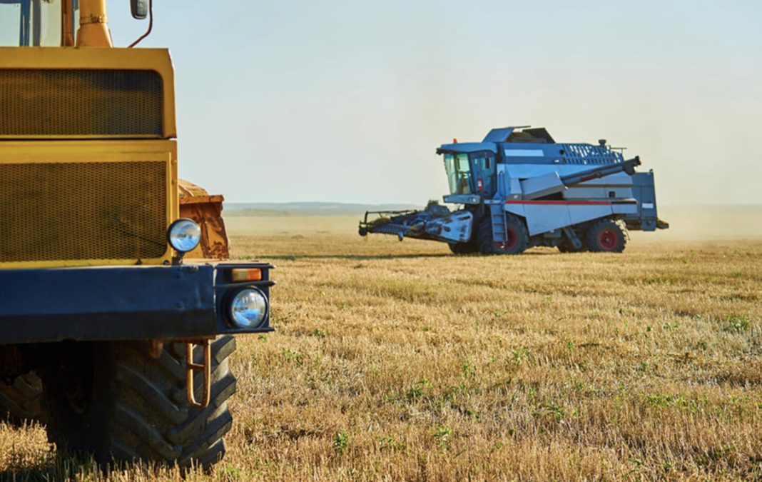 Two large agricultural machines in a dry field, a yellow vehicle in the foreground and a blue combine harvester in the distance under a clear sky.