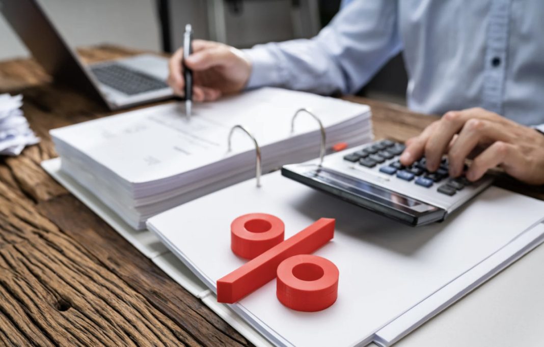 Person using a calculator and writing in a binder-filled notebook, with a large red percent sign on the desk (finance/accounting scene).