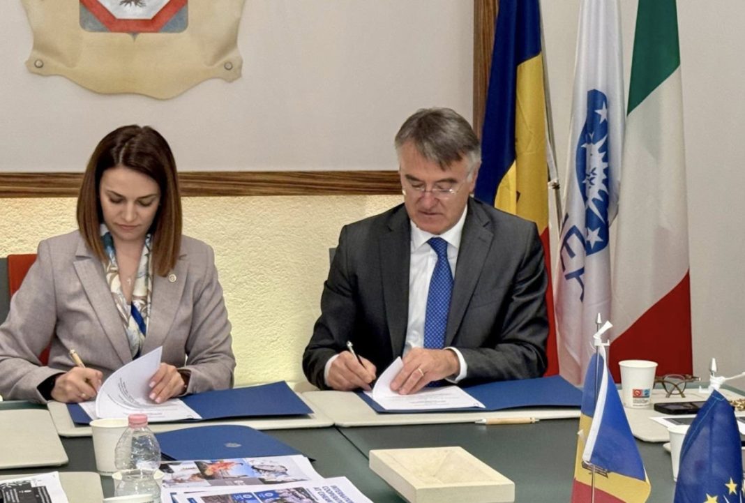 Two professionals sign documents at a formal meeting, a man in a suit and a woman in a blazer seated at a table with blue folders and flags in the background.