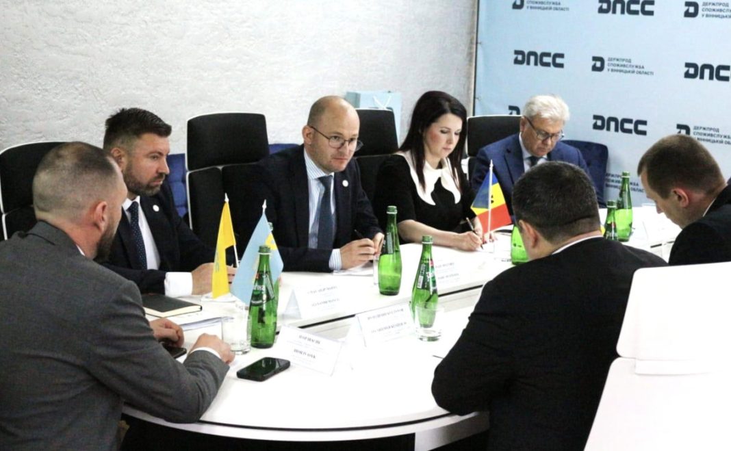 Formal meeting of officials around a round table with small flags, name cards, and green glass bottles in a conference room, DNCC backdrop visible.