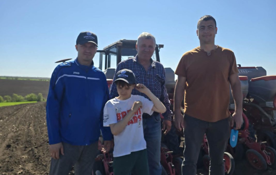 Four people pose in a plowed farm field with a tractor in the background.