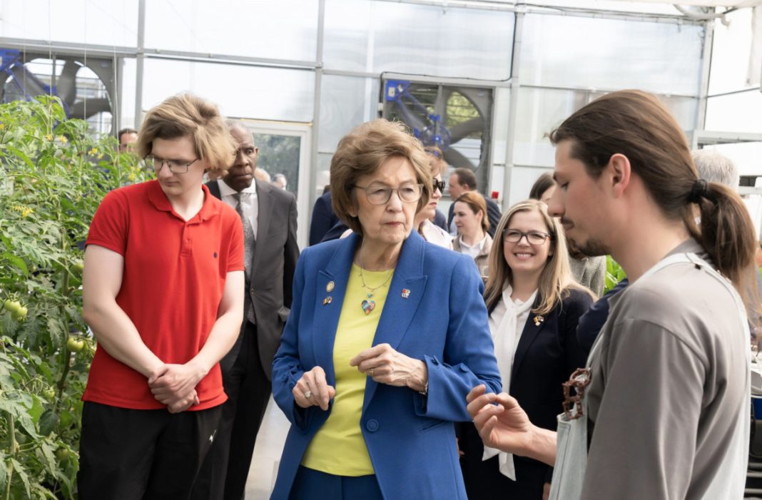 Group of people tour a greenhouse; a woman in a blue blazer talks with a young man in a gray shirt near tomato plants.