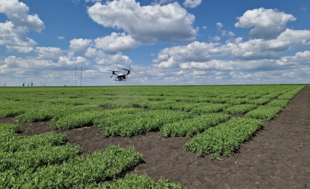 Drone flying over a patchwork farm with neat crop rows under a blue, cloud-filled sky. In the background, power lines trace the horizon.