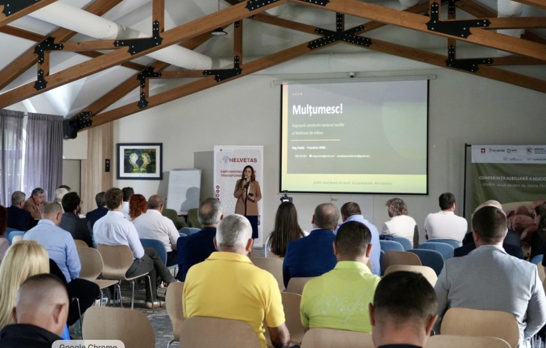 Woman presenting at a conference with a projector screen showing 'Mulțumesc!' in a room with wooden beam ceiling and an attentive audience.