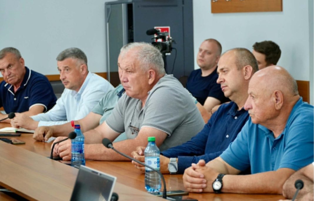 Group of men seated along a conference table in a meeting room, microphones in front, suggesting a formal briefing or discussion.
