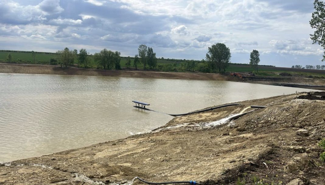 Muddy shoreline with a small blue dock extending into a calm body of water, construction equipment visible in the background under a cloudy sky.