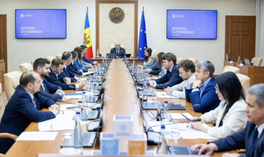 Large government meeting around a long wooden conference table, with officials on both sides, laptops and microphones, and a head of the table flanked by Moldova and EU flags behind him.