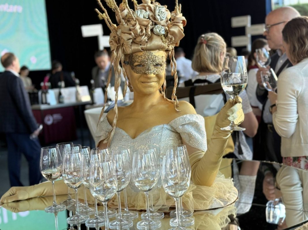 Person in gold body paint with an elaborate headpiece and mask holds up a wine glass at a reception while surrounded by empty glasses.]
