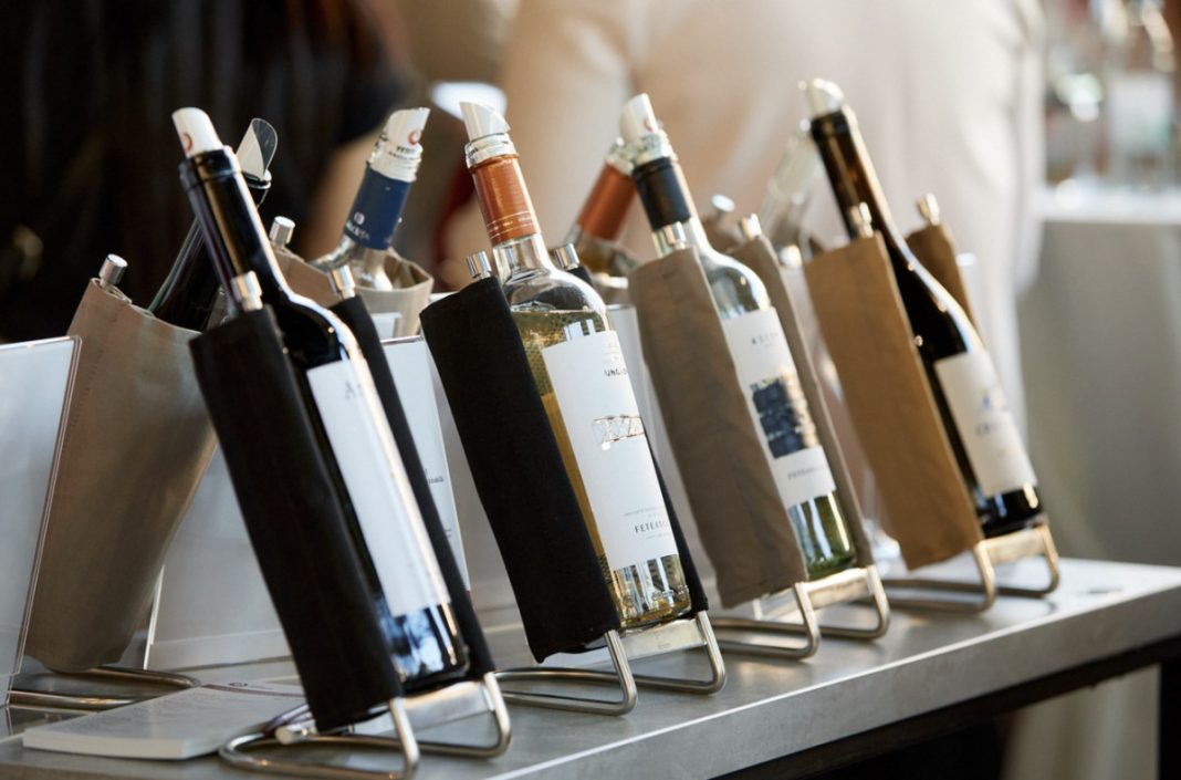Wine bottles displayed on angled metal racks at a bar or tasting counter, wrapped in paper sleeves for a selection event.