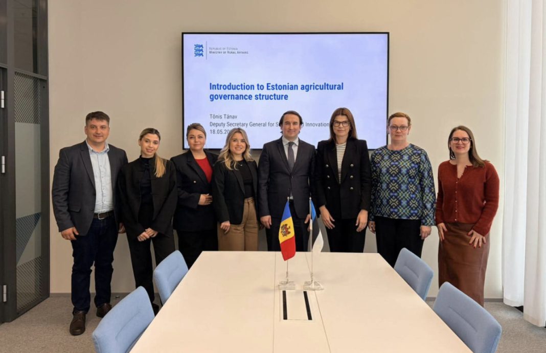 Group of eight professionals in business attire posing in a meeting room, with a large presentation screen behind them reading 'Introduction to Estonian agricultural governance structure' and two small flags on the table in front of them.