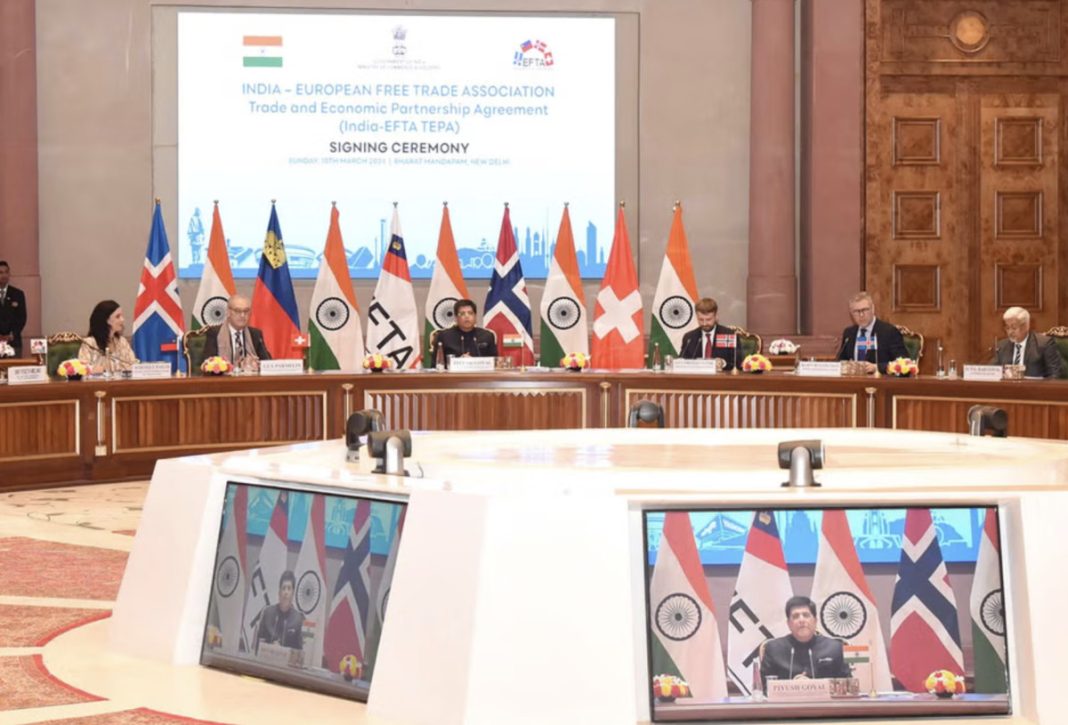 Signing ceremony for India–European Free Trade Association with flags and officials at a long table in a grand hall; large screen displays event title.