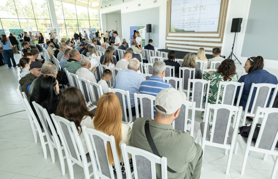 Audience seated in a bright conference hall, facing a stage with a large screen visible above the room.