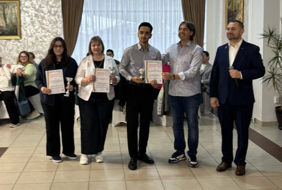 Five people pose with diplomas and trophies at an awards ceremony, the man on the right holding a microphone.