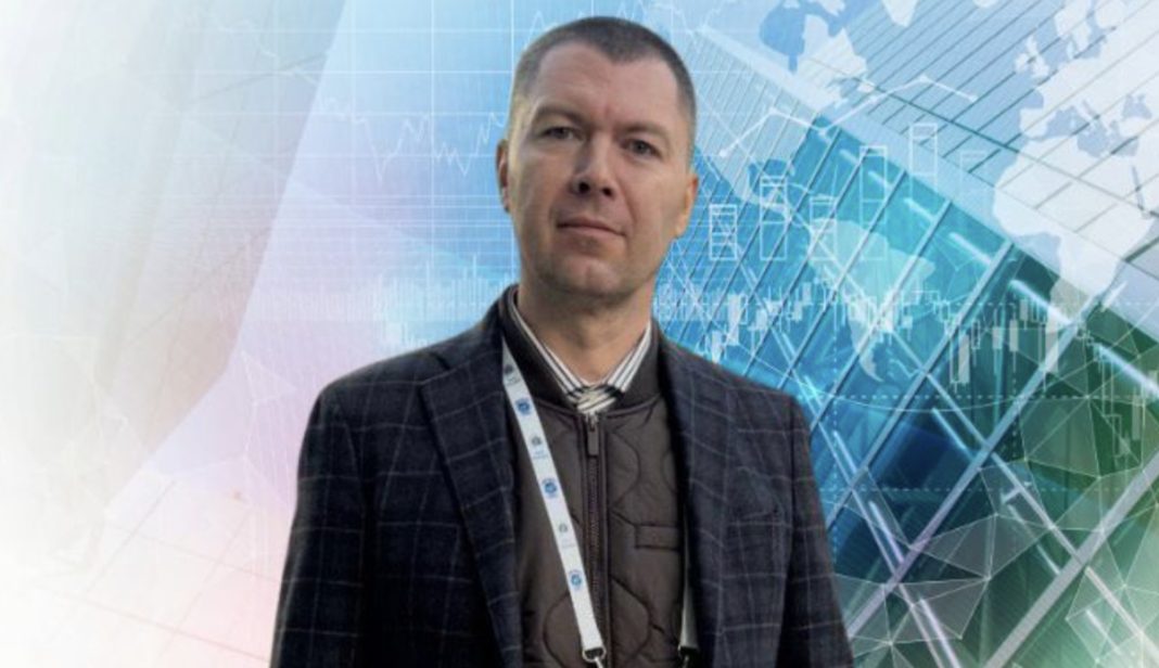 Man in a dark checkered blazer wearing a lanyard badge stands against a blue-green abstract tech background with graphs and grids.