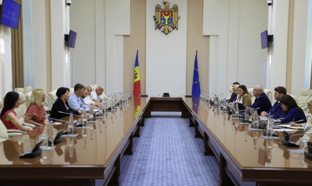 Two delegations sit on opposite sides of a long conference table in a government chamber, Moldova and EU flags behind them and a crest on the wall.