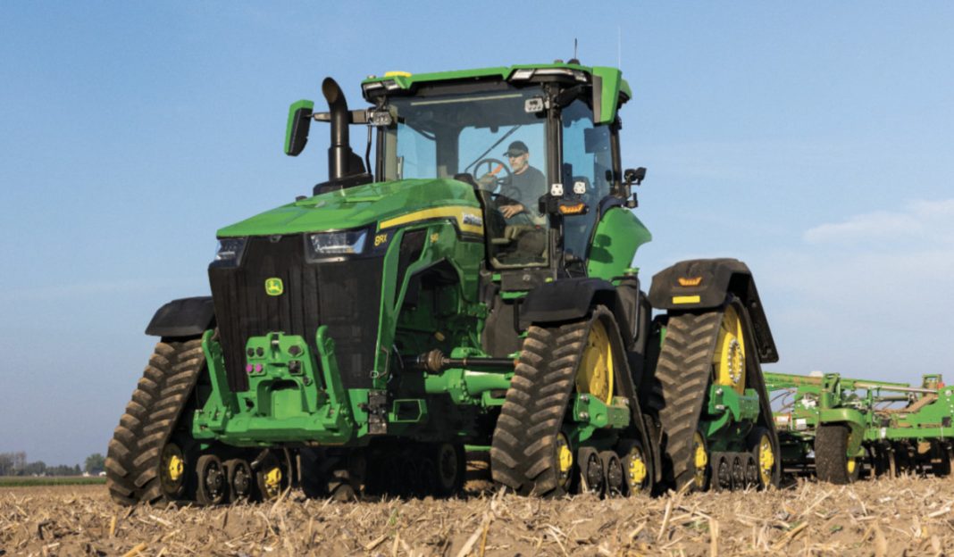 Green John Deere tracked tractor in a field with a farmer driving, pulling farming equipment under a clear blue sky