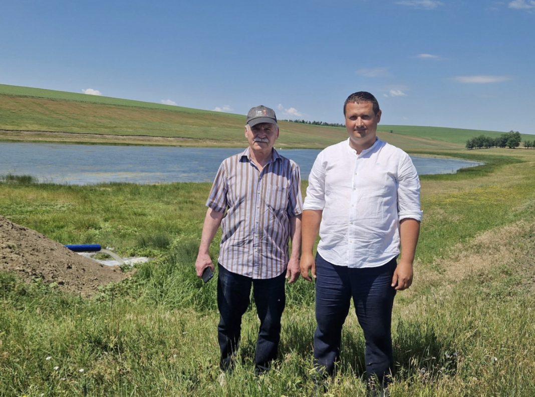 Two men stand on a grassy bank beside a small pond under a clear blue sky on a sunny day in the countryside.