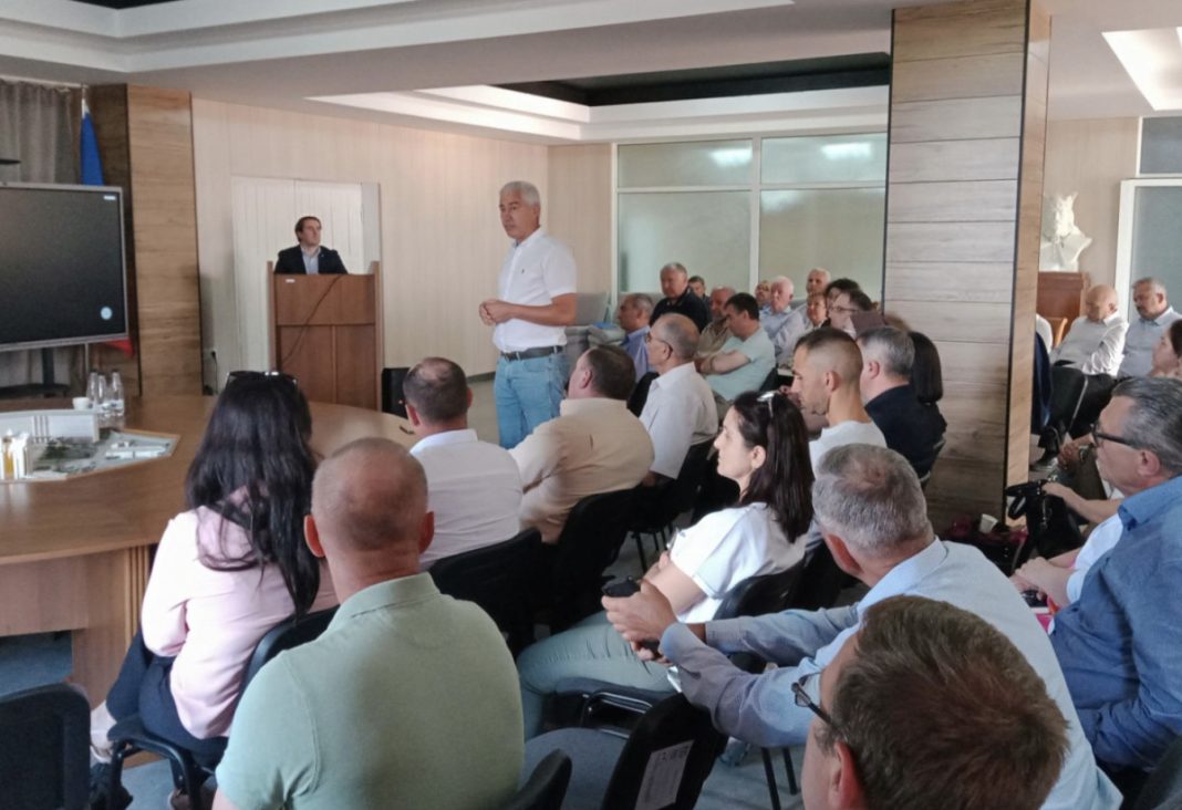 Man in a white shirt stands and speaks to seated attendees in a conference room.