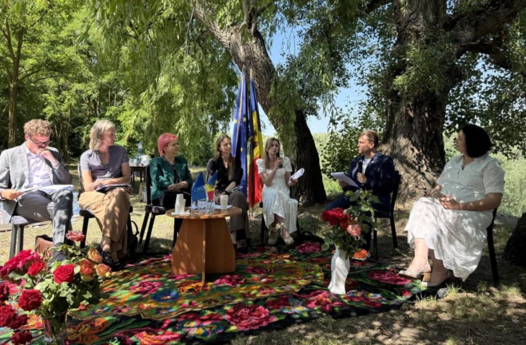 Outdoor panel discussion under trees with seven people seated in a semicircle; flags on a small table and a vibrant floral rug beneath them.