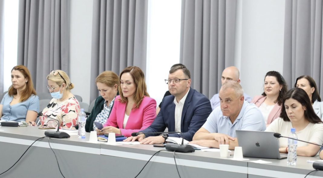 Group of professionals seated along a long conference table during a meeting, with notebooks, laptops, and microphones in front of them.
