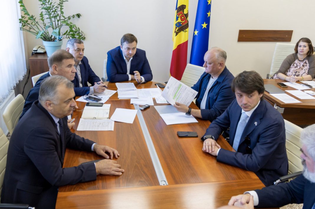 Group of professionals in suits sit around a wooden conference table discussing documents, with Moldova and EU flags in the background.