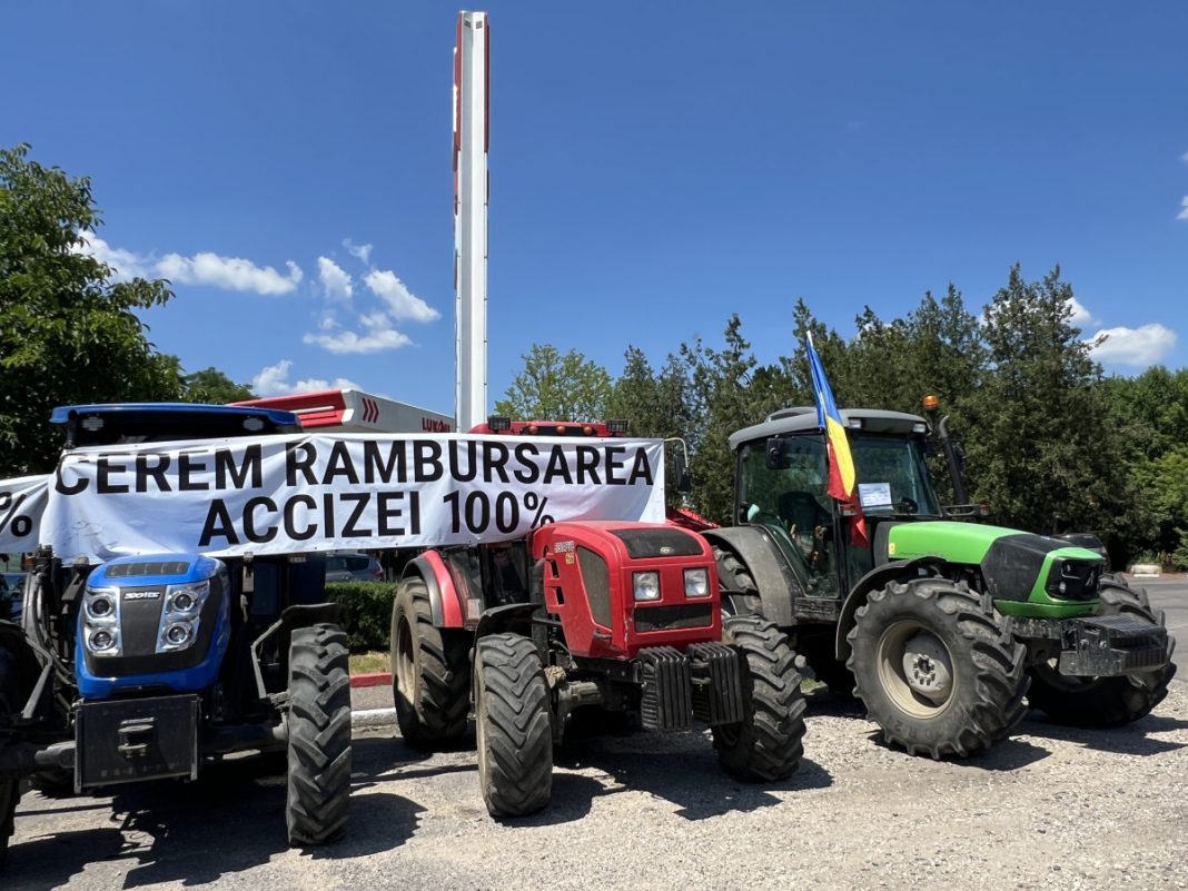 Line of farm tractors with a banner reading 'Cerem rambursarea accizei 100%' in a parking lot, a Romanian flag visible on the tractor site.