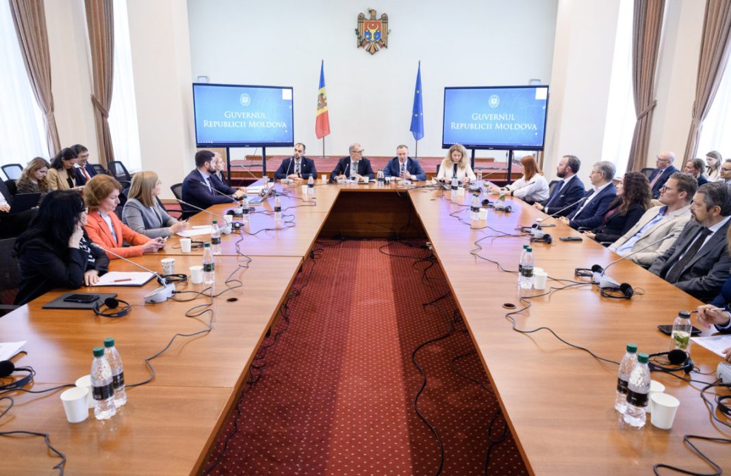 Government officials seated around a large U-shaped conference table in a formal meeting room with Moldova flags and emblem on the wall.