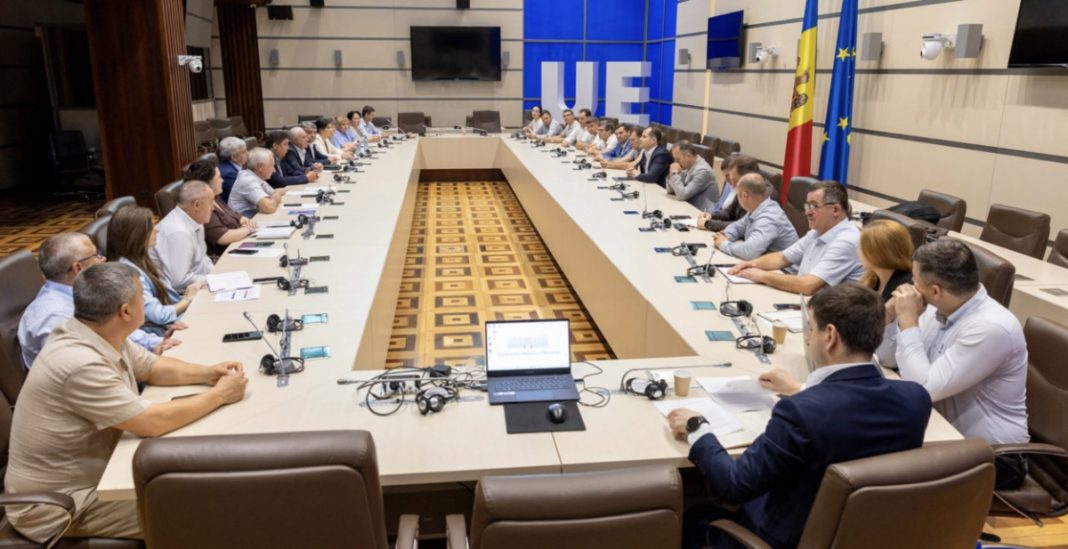 Large conference room with a long U-shaped table and many delegates in formal attire around it, microphones and laptops on the table, European flag in the room.