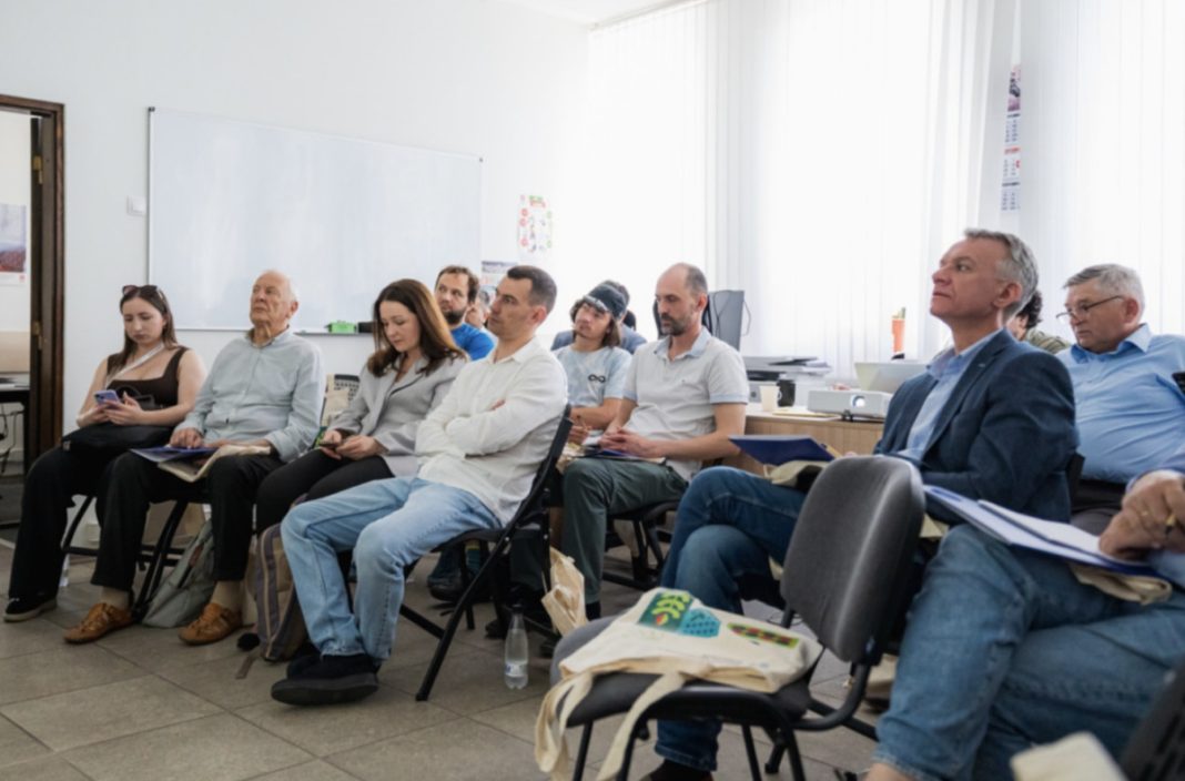 Group of adults seated in a classroom listening to a presentation near a whiteboard, some with notebooks and bags at their feet.