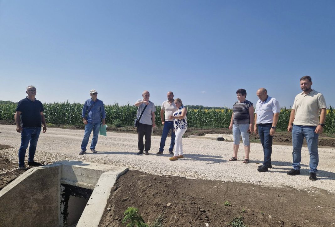 Group of people standing on a gravel road beside an open concrete drainage or construction pit, pointing and discussing on a sunny day with a cornfield in the background.