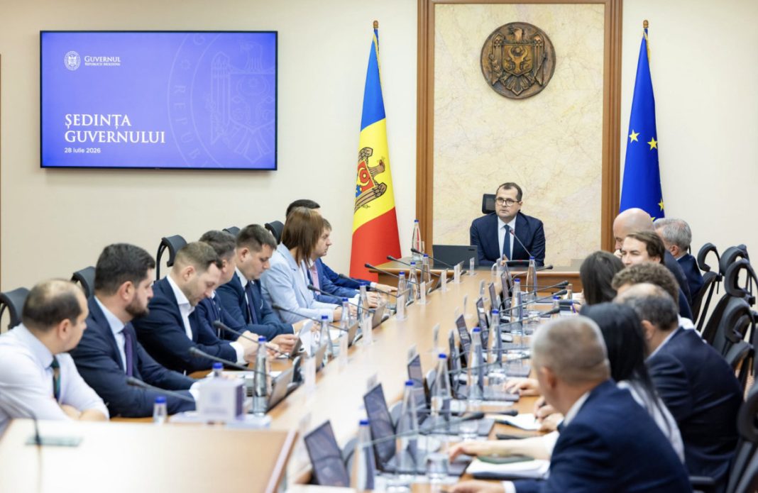 Government ministers sit around a long conference table in a formal meeting room with Moldova and EU flags; screen on wall reads 'ȘEDINȚA GUVERNULULUI 28 iulie 2026'
