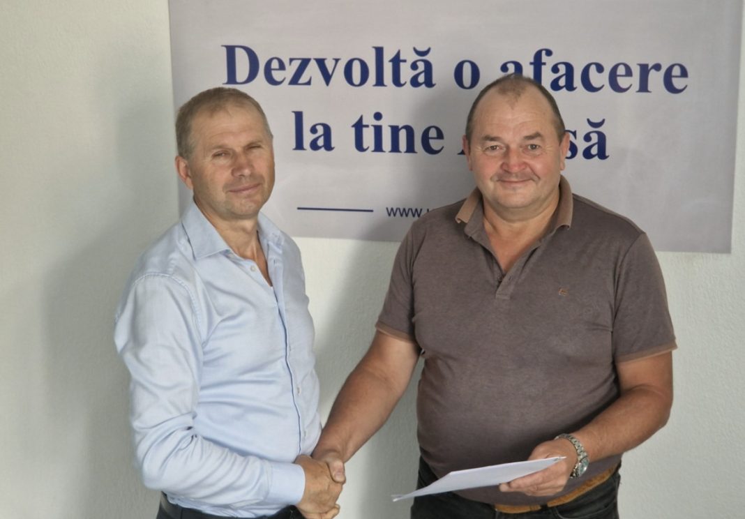 Two men shake hands in front of a sign, one holding a sheet of paper, suggesting a formal agreement.