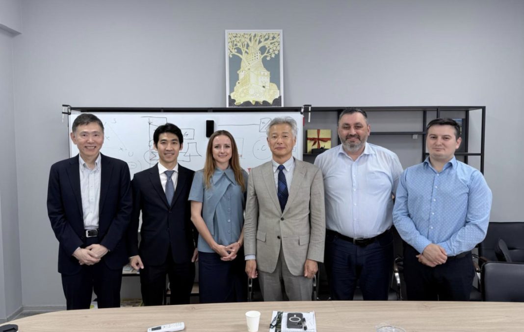 Six professionals in business attire pose for a group photo in an office, with a whiteboard and shelves in the background.