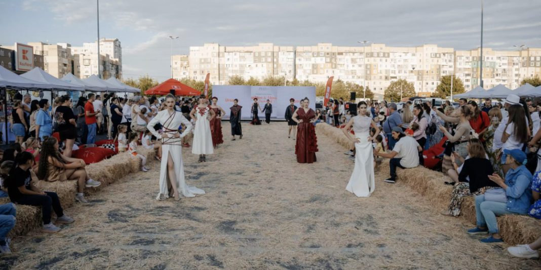 Outdoor fashion show on a hay runway with models in white and red gowns, audience seated on hay bales under tents.