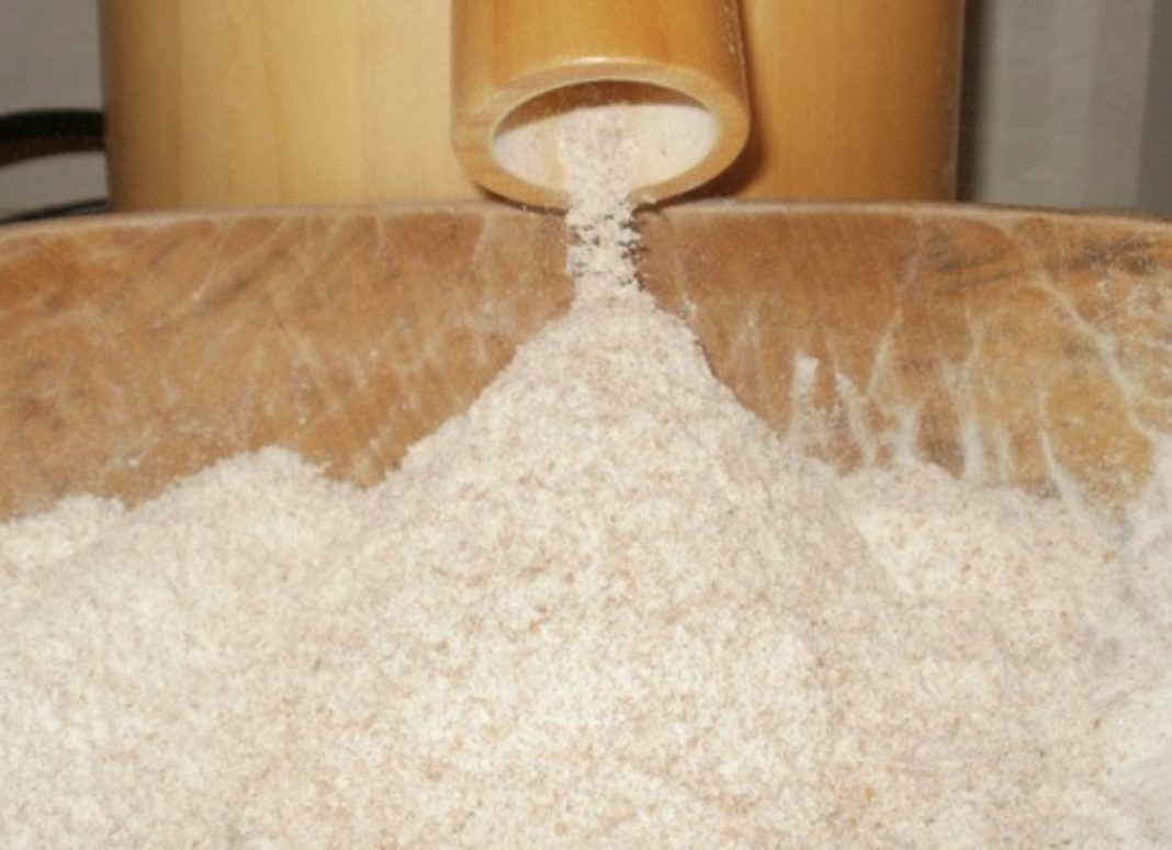 Wooden scoop pours white flour into a wooden trough or container.