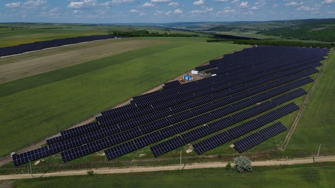 Aerial view of a large solar farm on a grassy hillside with rows of dark panels and a small construction area in the center.