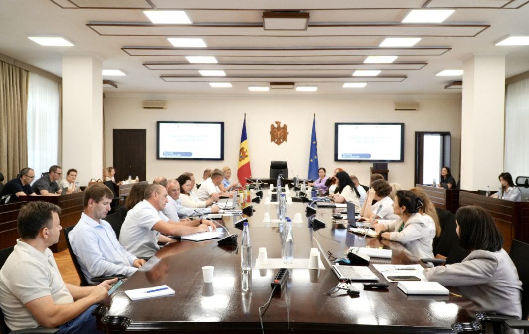 Formal boardroom meeting with a long dark wooden table, attendees around it, two presentation screens at the front, and flags behind the central seats.