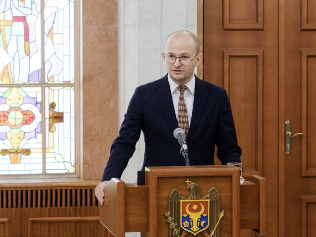 Man in a dark suit and tie speaks from a wooden podium in a room with a stained-glass window behind him.
