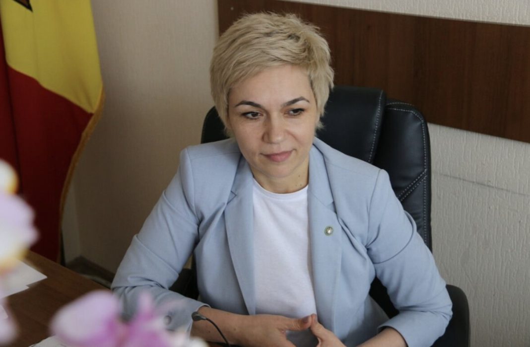 Woman with short blonde hair seated at a desk in a light blue blazer and white shirt, looking slightly to the side in an office with a flag behind her.