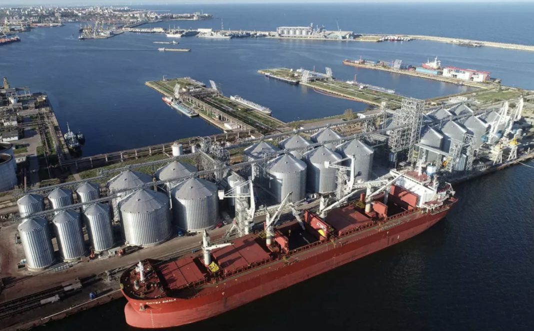 Aerial view of a port with a red cargo ship docked beside long rows of silver grain silos and cranes along the quay.
